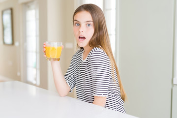 Beautiful young girl kid drinking a glass of fresh orange juice scared in shock with a surprise face, afraid and excited with fear expression