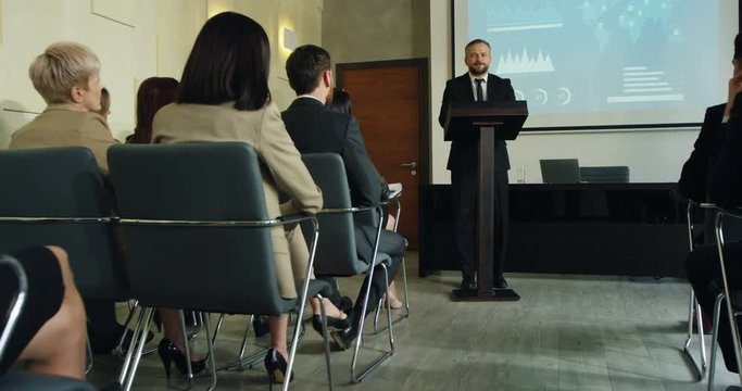 Camera zooming in on the Caucasian male business trainer having an educational lection in front of the many people and demonstrating some charts and graphics on the big screen.
