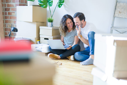 Middle age senior romantic couple in love sitting on the apartment floor with boxes around and using computer laptop smiling happy for moving to a new home