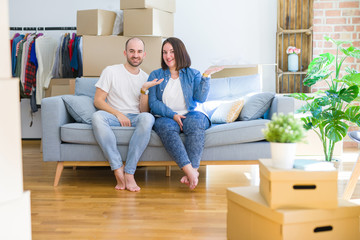 Fototapeta na wymiar Young couple sitting on the sofa arround cardboard boxes moving to a new house smiling cheerful presenting and pointing with palm of hand looking at the camera.