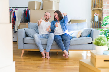 Fototapeta na wymiar Young couple sitting on the sofa arround cardboard boxes moving to a new house approving doing positive gesture with hand, thumbs up smiling and happy for success. Winner gesture.