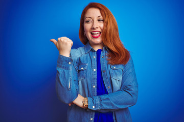 Young beautiful redhead woman wearing denim shirt standing over blue isolated background smiling with happy face looking and pointing to the side with thumb up.