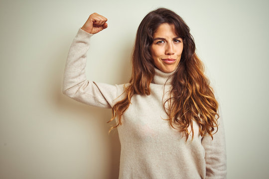 Young beautiful woman wearing winter sweater standing over white isolated background Strong person showing arm muscle, confident and proud of power
