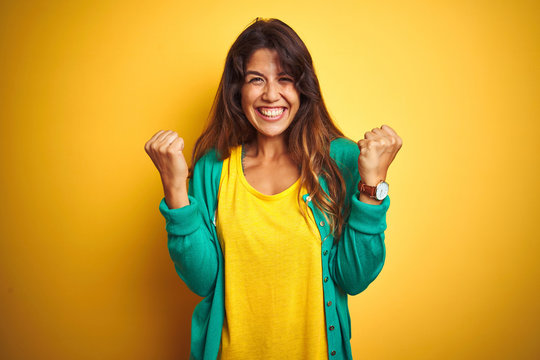Young woman wearing t-shirt and green sweater standing over yelllow isolated background celebrating surprised and amazed for success with arms raised and open eyes. Winner concept.