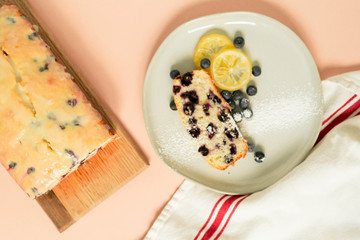 Slice of Iced Lemon Blueberry Loaf Cake on Plate with Lemons and Blueberries, Pink / Coral Background, Top View / Flat Lay