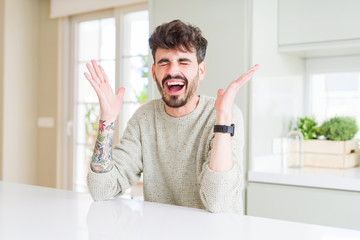 Young man wearing casual sweater sitting on white table celebrating mad and crazy for success with...