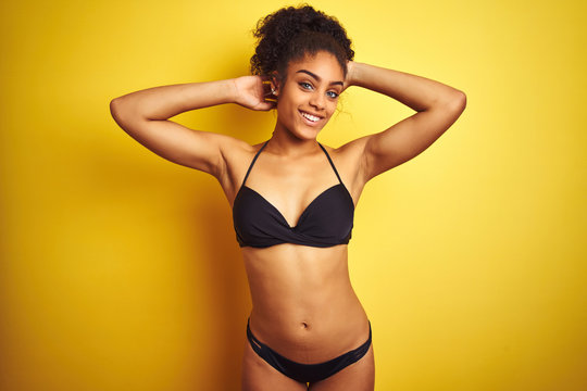African american woman on vacation wearing bikini standing over isolated yellow background relaxing and stretching, arms and hands behind head and neck smiling happy