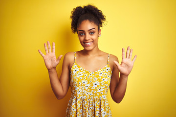 African american woman wearing casual floral dress standing over isolated yellow background showing and pointing up with fingers number ten while smiling confident and happy.