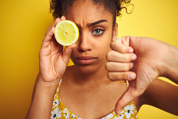 Young african american woman holding slice of lemon over isolated yellow background with angry...