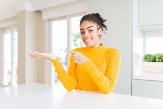 Beautiful african american woman with afro hair wearing a casual yellow sweater amazed and smiling to the camera while presenting with hand and pointing with finger.