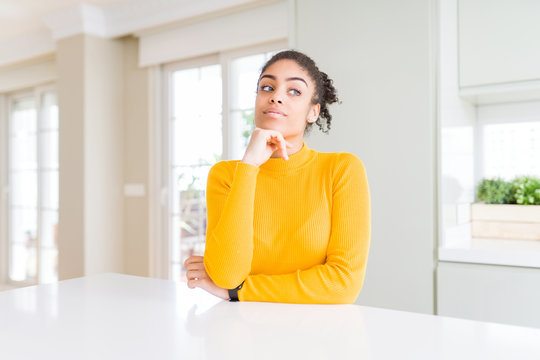 Beautiful african american woman with afro hair wearing a casual yellow sweater with hand on chin thinking about question, pensive expression. Smiling with thoughtful face. Doubt concept.