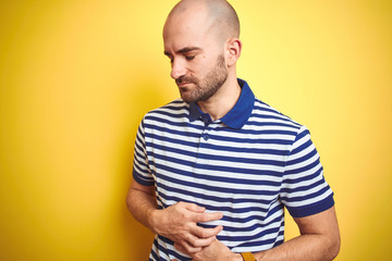 Young bald man with beard wearing casual striped blue t-shirt over yellow isolated background with hand on stomach because nausea, painful disease feeling unwell. Ache concept.