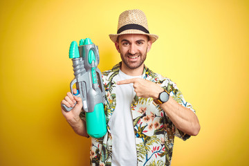 Young man wearing hawaiian flowers shirt holding water gun over yellow isolated background very happy pointing with hand and finger