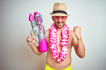 Young man wearing summer hat and hawaiian lei flowers holding water gun over isolated background...