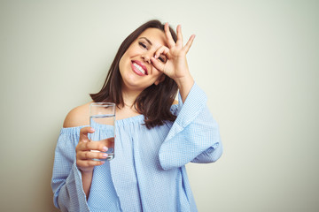 Young beautiful woman drinking a glass of fresh water over isolated background with happy face smiling doing ok sign with hand on eye looking through fingers