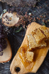 A table with a wicker table top, honey in honeycombs on a wooden board, honey flows from a spoon into an open glass jar. Medicinal herbs oregano, chamomile and strawberry flowers. Flower pollen collec