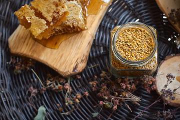 A table with a wicker table top, honey in honeycombs on a wooden board, honey in an open glass jar, with medicinal herbs oregano, chamomile and strawberry flowers. flower pollen collected by bees