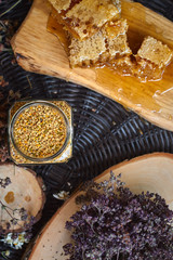 A table with a wicker table top, honey in honeycombs on a wooden board, honey in an open glass jar, with medicinal herbs oregano, chamomile and strawberry flowers. flower pollen collected by bees
