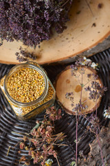 A table with a wicker table top, honey in honeycombs on a wooden board, honey in an open glass jar, with medicinal herbs oregano, chamomile and strawberry flowers. flower pollen collected by bees