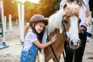 Cute little girl and her older sister enjoying with pony horse outdoors at ranch. - obrazy, fototapety, plakaty