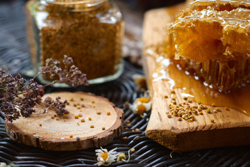 A table with a wicker table top, honey in honeycombs on a wooden board, honey in an open glass jar, with medicinal herbs oregano, chamomile and strawberry flowers. flower pollen collected by bees