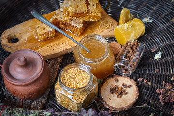 A table with a wicker table top, honey in honeycombs on a wooden board, honey in an open glass jar, with medicinal herbs oregano, chamomile and strawberry flowers. flower pollen collected by bees