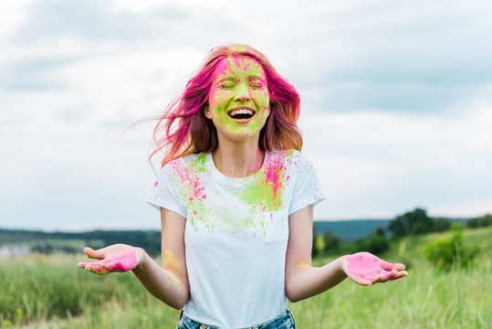 cheerful woman with closed eyes and pink holi paint on face gesturing and smiling outdoors