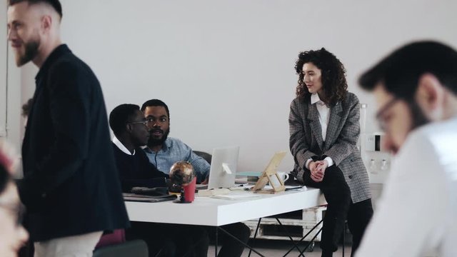 Wide shot, happy young boss business woman talking to two African male colleagues at busy modern office workplace table.