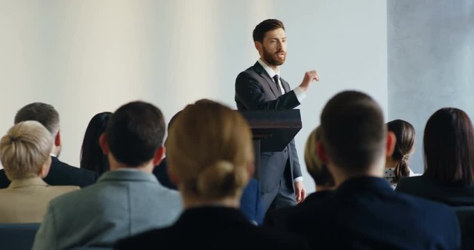 Young Caucasian business trainer walking at the podium and teaching his audience during the conference speech.