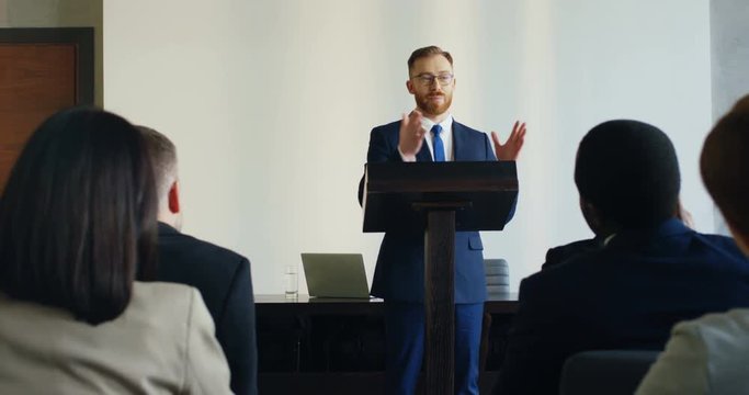 View from among the people in audience on the Caucasian god looking male business coach standing at the lecturn and speaking to the audience.