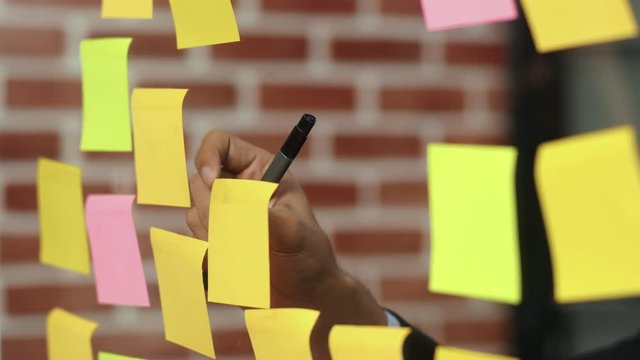 Asian creative man stick a sticky note on glass board. Young professional business male write information, strategy, reminder on paper, business situation, startup in Loft office concept. Slow motion.