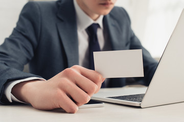 partial view of businessman showing blank business card at camera while using laptop