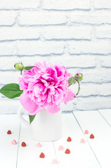 pink peony flower in white vase with red and pink hearts on brick wall background.