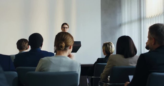 Young beautiful Caucasian woman professor standing at the lecturn and telling or explaining something to the people who sitting in front of her.