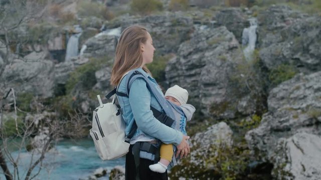 Young mother with her son in ergo backpack travels in nature near the waterfall in spring, Montenegro