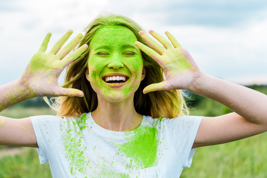 cheerful woman with closed eyes and green holi paint on hands gesturing and smiling outdoors