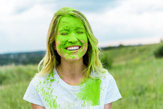 cheerful woman with closed eyes and green holi paint on face smiling outdoors