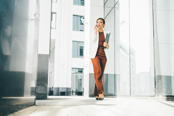 Successful businesswoman talking on phone while walking outdoor. Reflecting in modern white bilding.