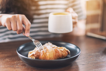 Closeup image of a woman eating a piece of croissant by fork for breakfast with coffee cup on wooden table