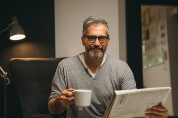 middle aged bearded man reading newspaper and having cup of coffee in his home
