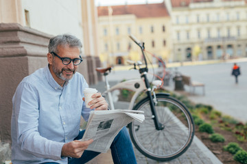 middle aged man reading newspaper and drinking coffee outdoor in town