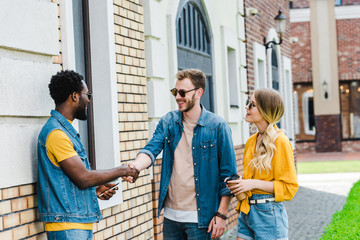 cheerful multicultural men shaking hands near happy woman with paper cup