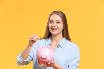 Fototapeta na wymiar Young woman putting coin into piggy bank on color background