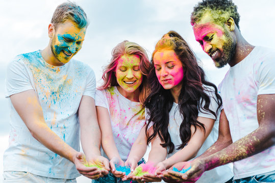 happy girls near multicultural men holding holi paints