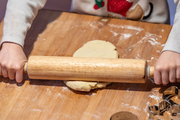 Kids hands rolling a piece of a cookie dough into a a thin pastry sheet using wooden roller pin.