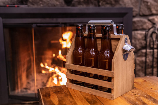 Handmade wooden carrying case for six beer bottles with a metal bottle opener on a side against a flame in a fireplace