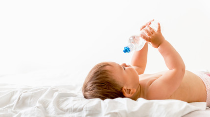 Baby toddler sits on the white bed, smiles and drinks water from plastic bottle. Copy space