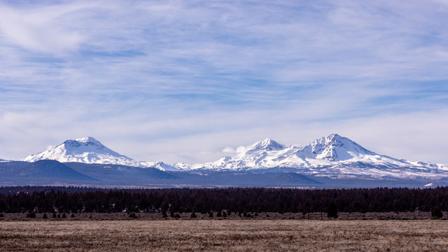 Three Sisters - panoramic view at mountains in Central Oregon with blue sky , white clouds and brown fields on a foreground