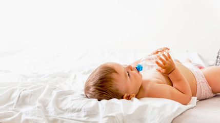 Baby toddler sits on the white bed, smiles and drinks water from plastic bottle. Copy space