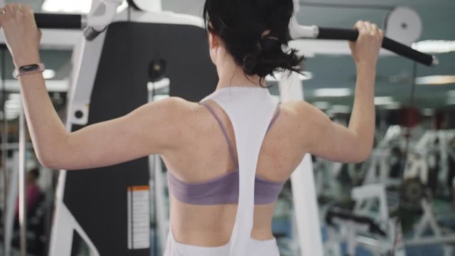 A woman does the exercises correctly, pulling up with a straight back. Fitness coach of the sports club does pull-ups on the simulator for strengthening the shoulder muscles.